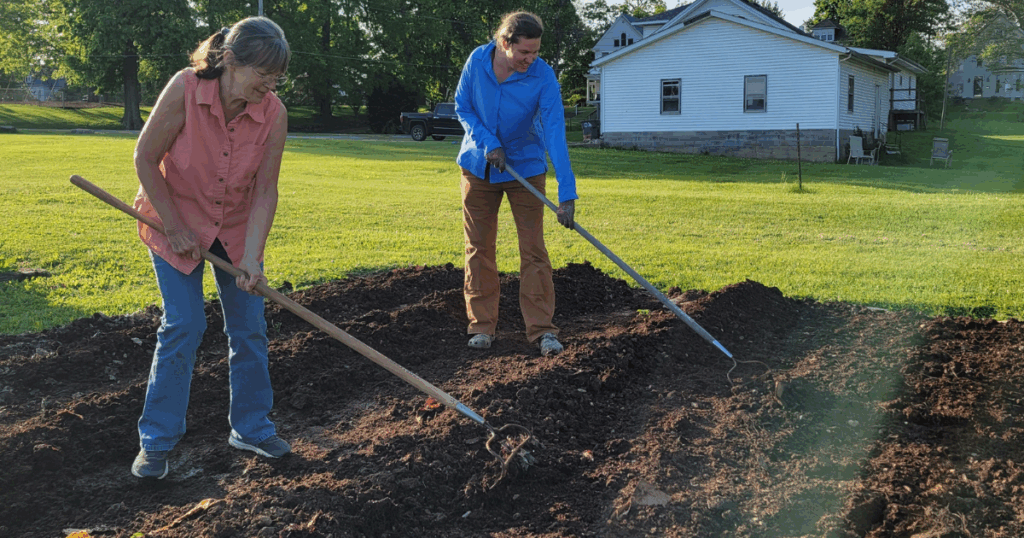 Two women using hoes to prepare soil for a garden.